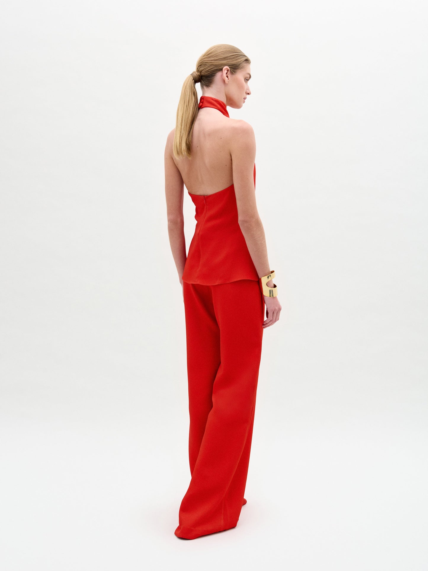 A woman poses against a white background in the sleeveless red halter top and high-waisted, wide-leg Palermo Pant in hammered satin, accessorized with gold bracelets.