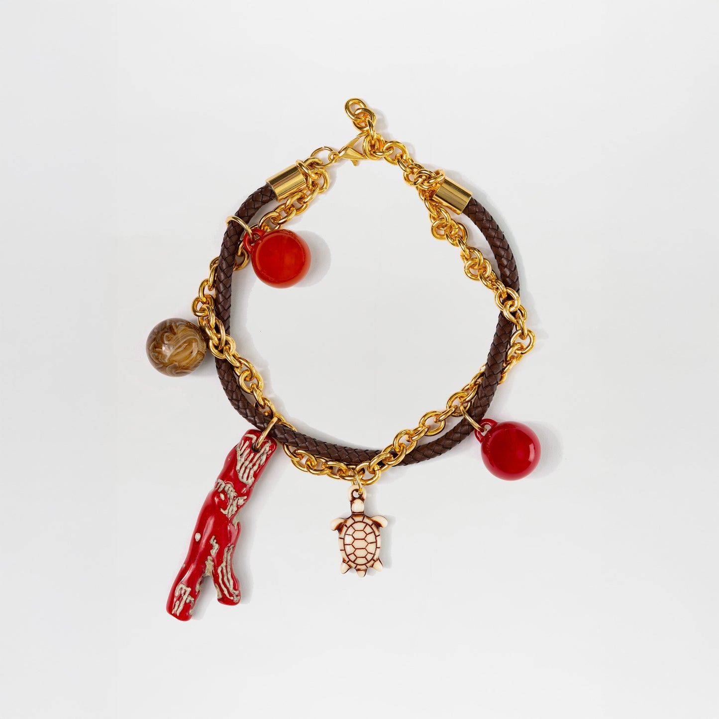 Collar Eira: cadena dorada con banda trenzada marrón, dije de tortuga dorado, abalorios rojos y marrones, charm de coral rojo sobre fondo blanco. Un diseño único con charms multicolores.