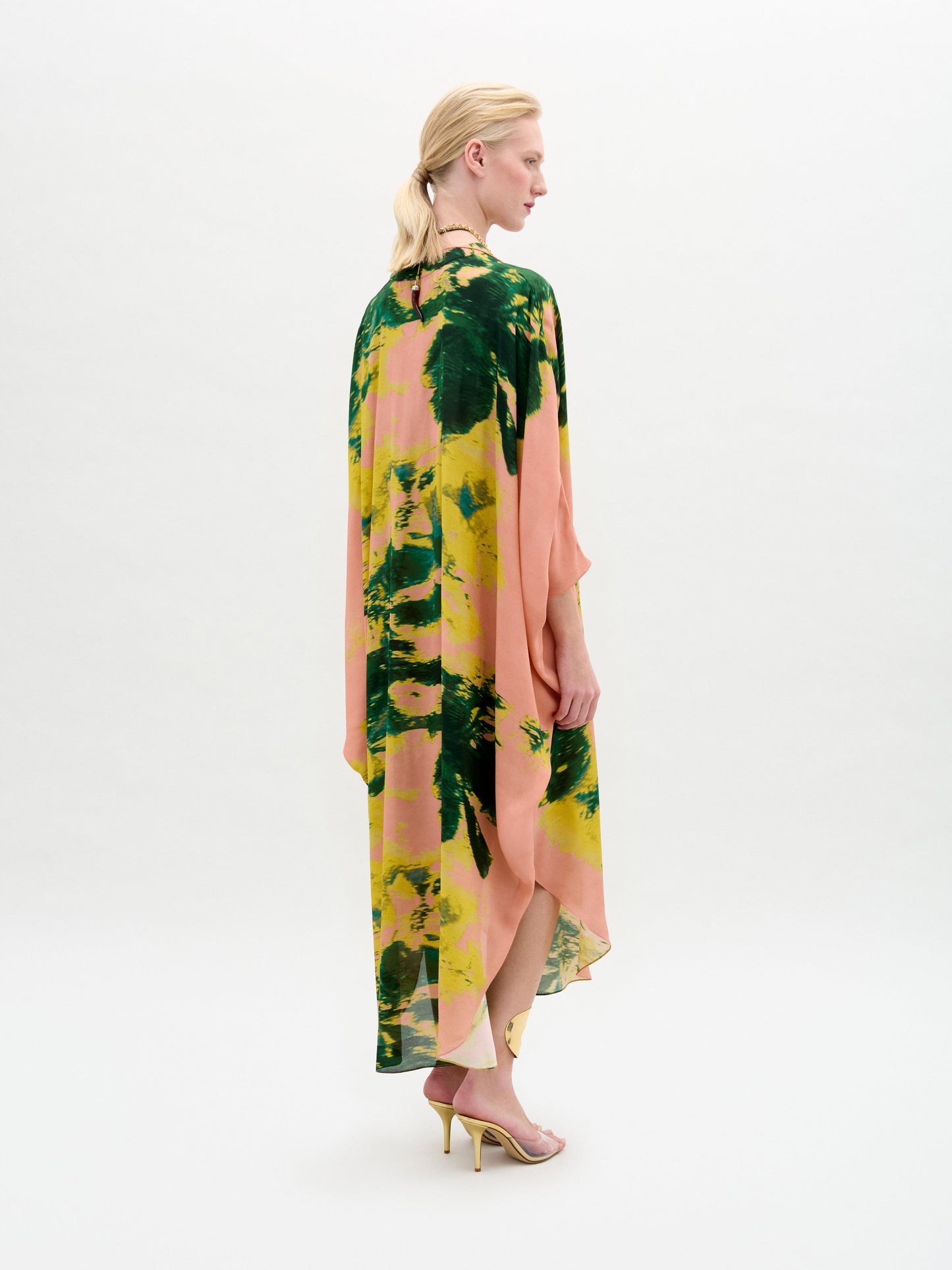 A woman stands against a white background wearing the Elea Tunic, an oversized 100% silk maxi dress with pink, yellow, and green patterns, paired with a beige bag and strappy heels.