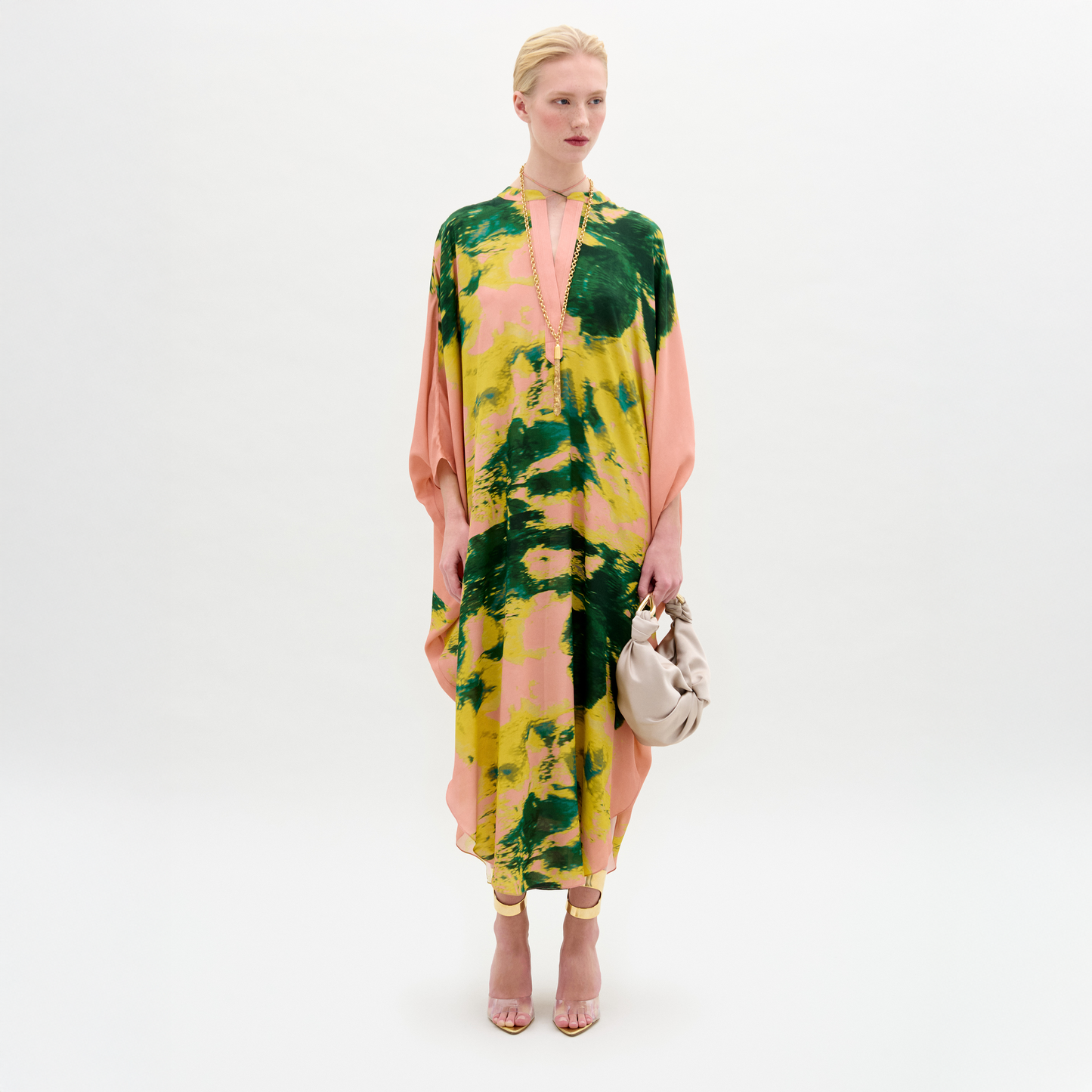 A woman stands against a white background wearing the Elea Tunic, an oversized 100% silk maxi dress with pink, yellow, and green patterns, paired with a beige bag and strappy heels.