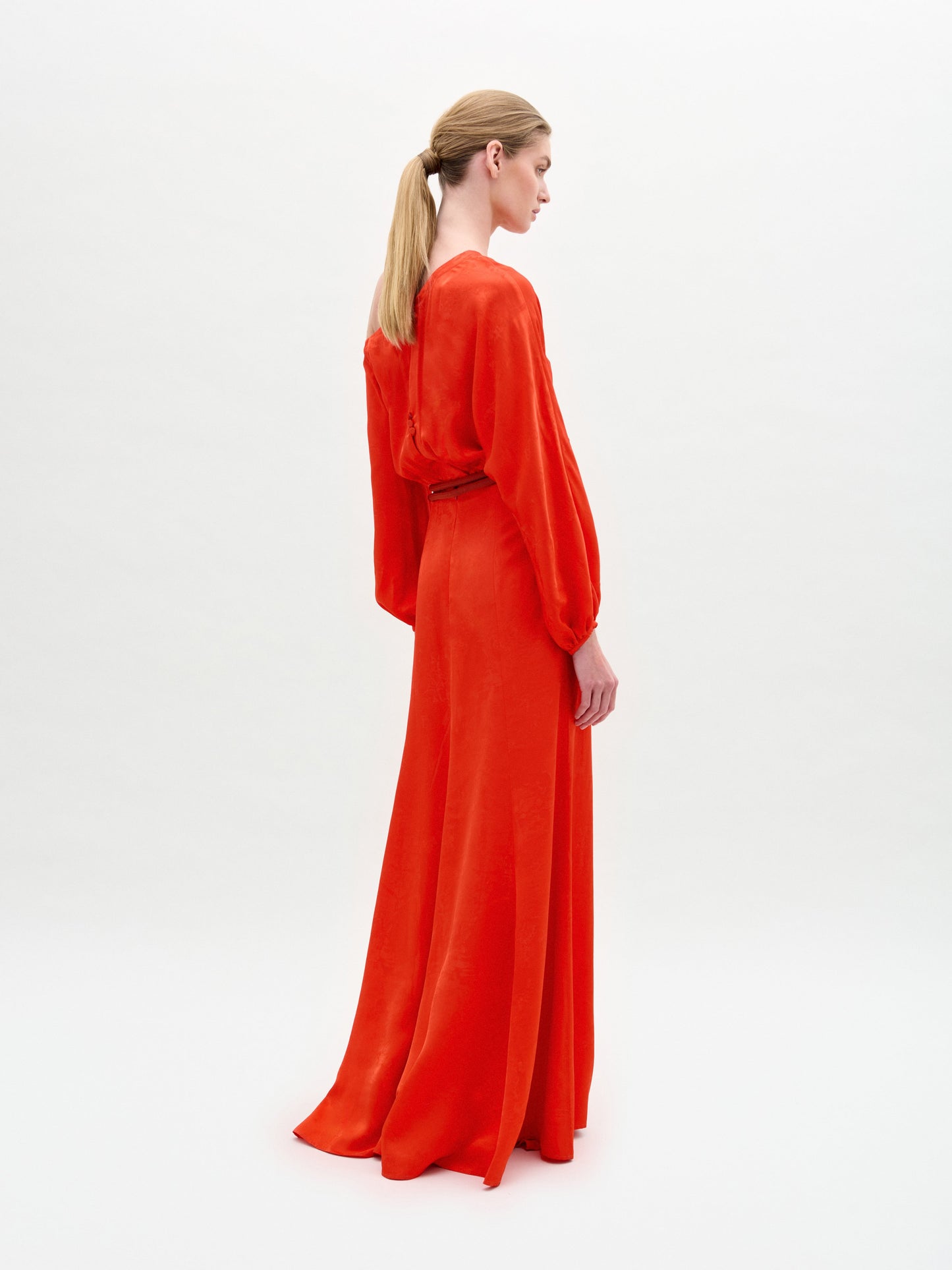 A woman poses against a white background in the Elga Dress—a red, floor-length, one-shoulder maxi dress with long sleeves and a gold 3D cord belt.