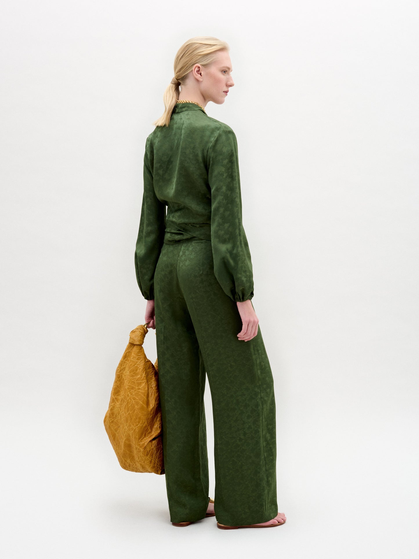 A woman stands against a plain background wearing wide-leg Emine Pants paired with a green Italian fabric blouse, holding a large textured mustard-yellow bag.