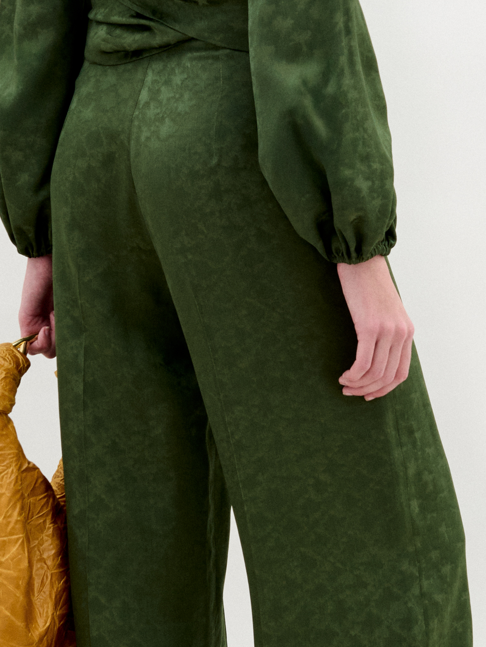 A woman stands against a plain background wearing wide-leg Emine Pants paired with a green Italian fabric blouse, holding a large textured mustard-yellow bag.
