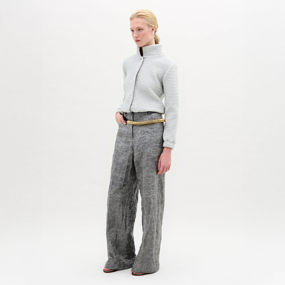A person stands in a white textured zip-up jacket and gray Emine Pant with a gold belt against a plain white background.
