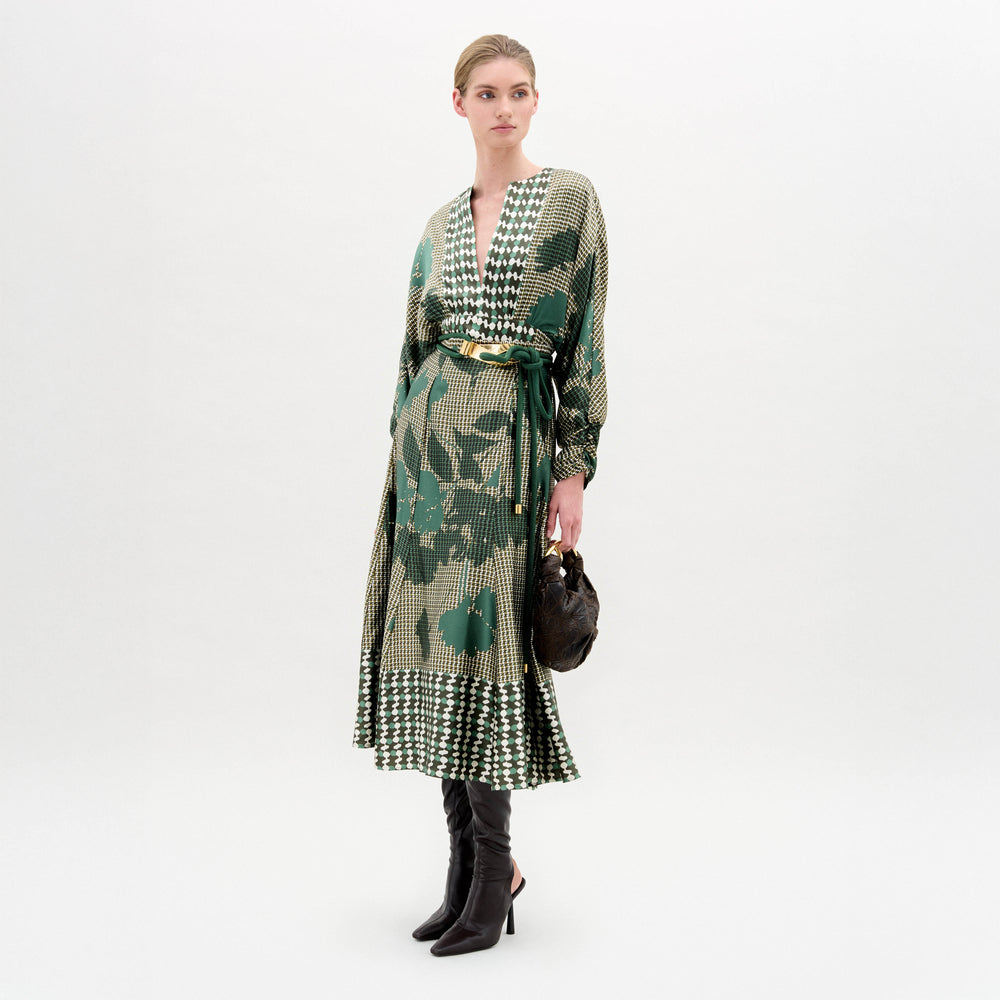 A woman stands against a white background wearing the Erin Skirt—a long, green and beige patterned skirt crafted from Italian fabric—paired with black heeled boots and a small black handbag.