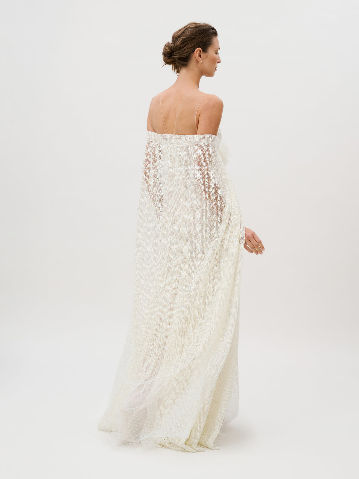 A woman stands against a white background, wearing the floor-length, off-the-shoulder Edna Dress made from sheer white fabric. Her hair is styled in a low, neat bun, and she is turned slightly to the side, facing away from the camera.