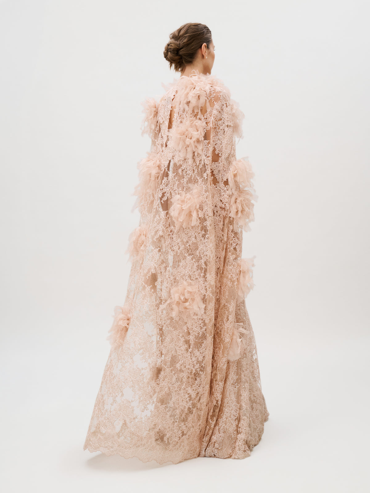 A woman in the Eudora Dress—a floor-length, pale pink lace gown adorned with floral appliqué and crafted from luxurious Italian fabric—stands against a plain white background, facing away from the camera with her hair styled in a low bun.