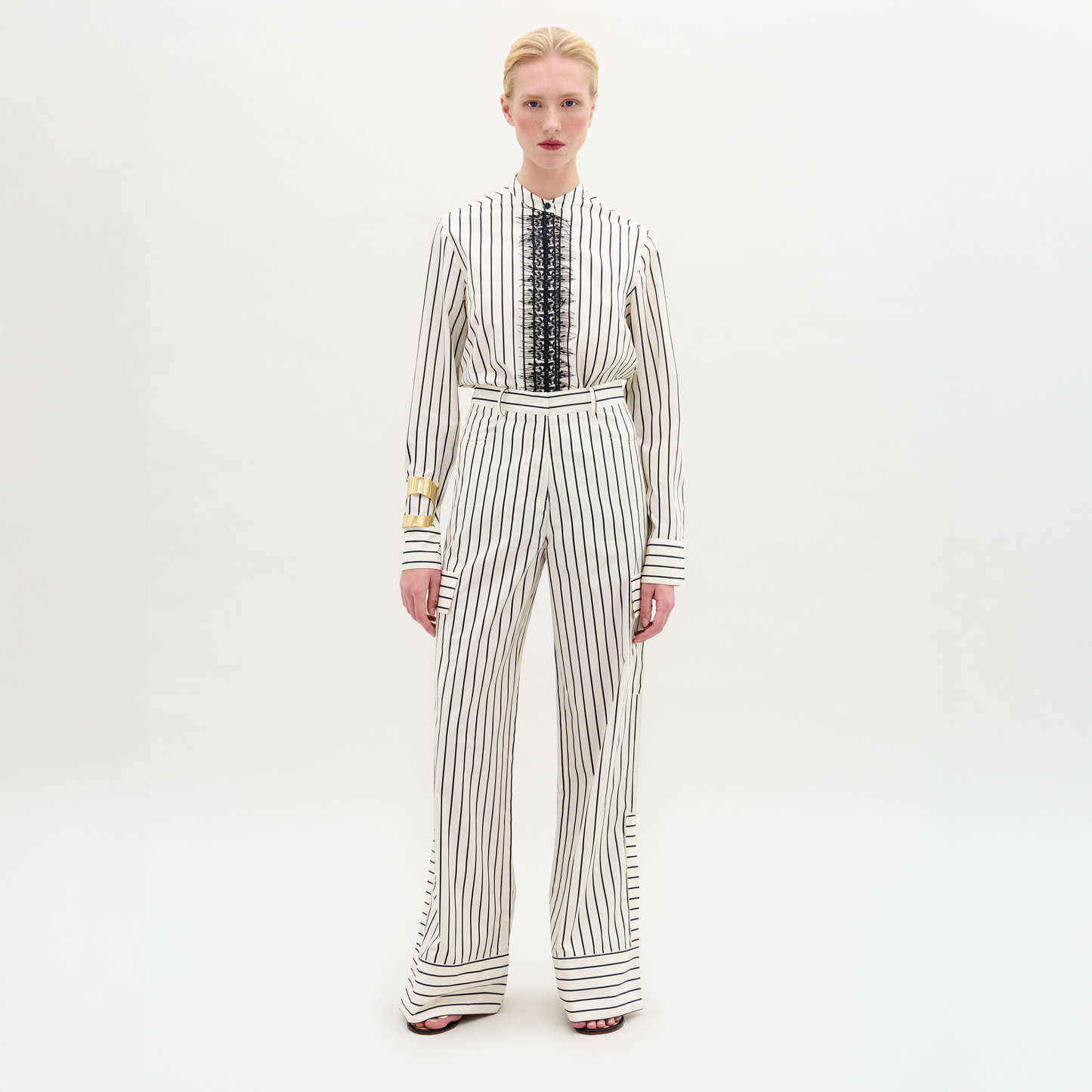 A person stands against a white background wearing a white long-sleeved blouse with vertical black stripes and lace detail, paired with the Falak Pant—relaxed fit, wide-leg trousers crafted from premium Italian fabric.