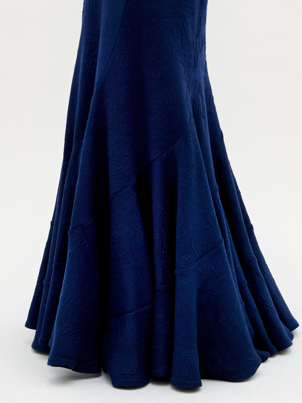 A woman wearing the Florina Skirt—a floor-length navy blue skirt with a mermaid silhouette and gathered knot detail at the waist—paired with gold jewelry, stands against a plain white background.