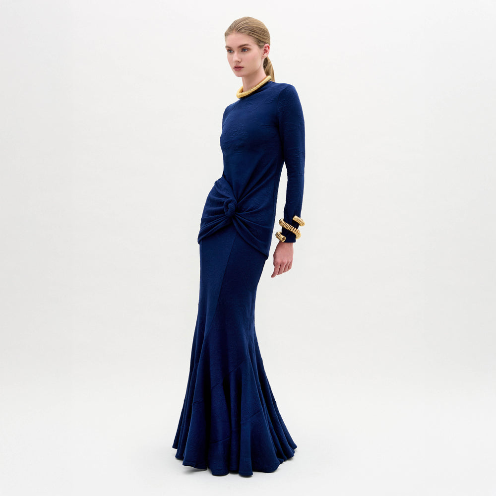 A woman wearing the Florina Skirt—a floor-length navy blue skirt with a mermaid silhouette and gathered knot detail at the waist—paired with gold jewelry, stands against a plain white background.