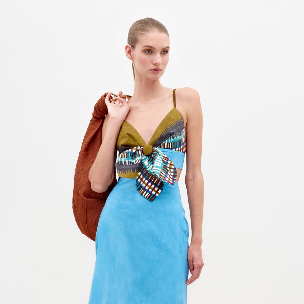A woman in an Aqua Olive Plaid Flor Top, featuring a colorful bow on the front, holds a brown jacket over her shoulder, ready for pre-order by February 15th, 2025.