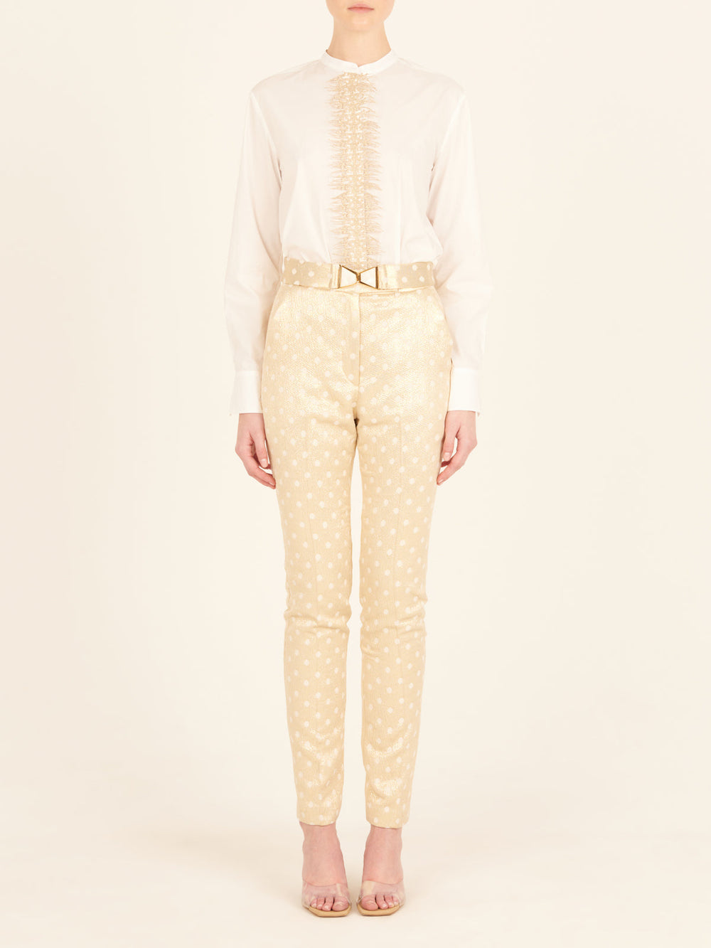 Rear view of a person with long hair in a ponytail, wearing a white blouse and the Orion Pant Gold Inverted, featuring high-waisted trousers with a straight leg silhouette, standing in heels on a plain background.