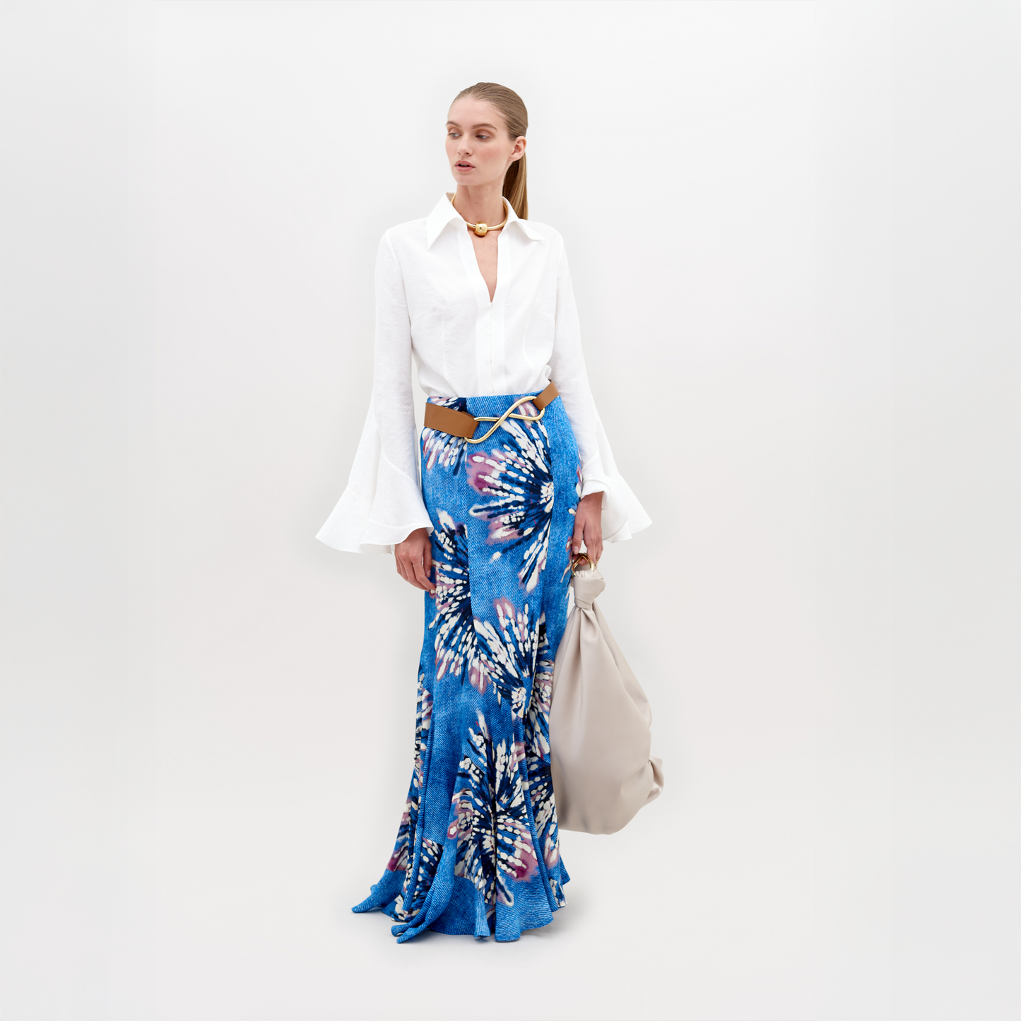 A woman in a white blouse with bell sleeves and a Florina Skirt Aciano Flower stands against a plain background, holding a light-colored oversized bag, available for pre-order. Mark your calendars: it will ship by November 15th, 2024.