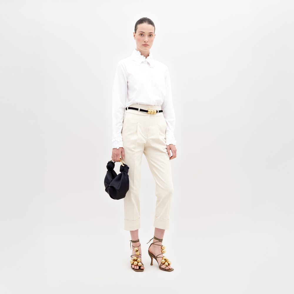 A person stands against a plain background, wearing a white blouse, Beryl Pant Beige, black belt, and golden embellished heels, holding a black handbag with a knot detail. This stylish ensemble is perfect for those who pre-order now to ensure it arrives by the November 15th, 2024 ship-by date.