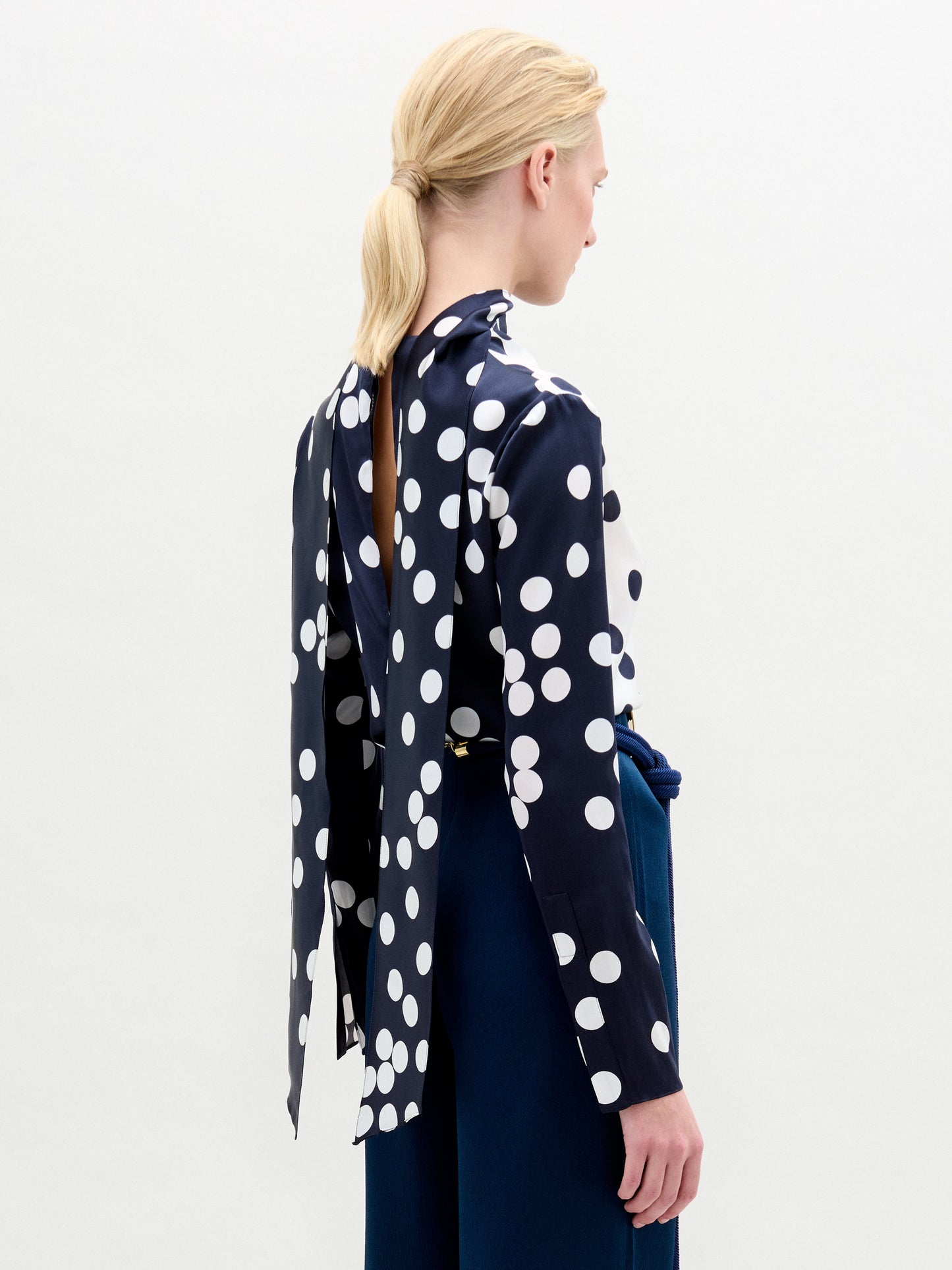A woman with pale skin and blonde hair wears the Gaby Blouse—a navy and white polka dot silk top with a keyhole neckline—paired with blue pants and a large circular belt buckle, standing against a plain background.