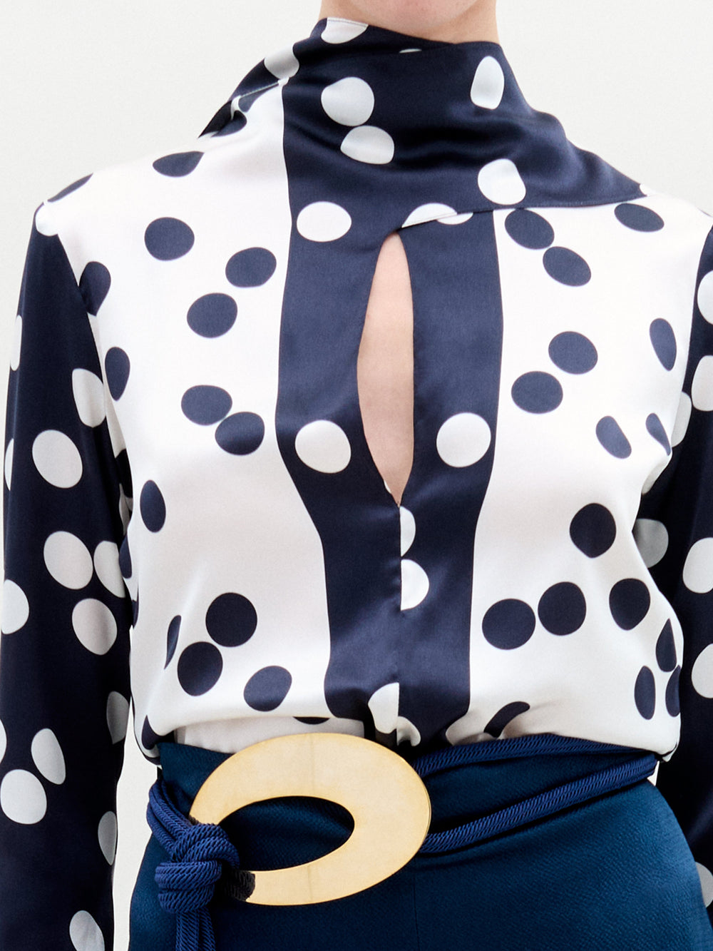 A woman with pale skin and blonde hair wears the Gaby Blouse—a navy and white polka dot silk top with a keyhole neckline—paired with blue pants and a large circular belt buckle, standing against a plain background.