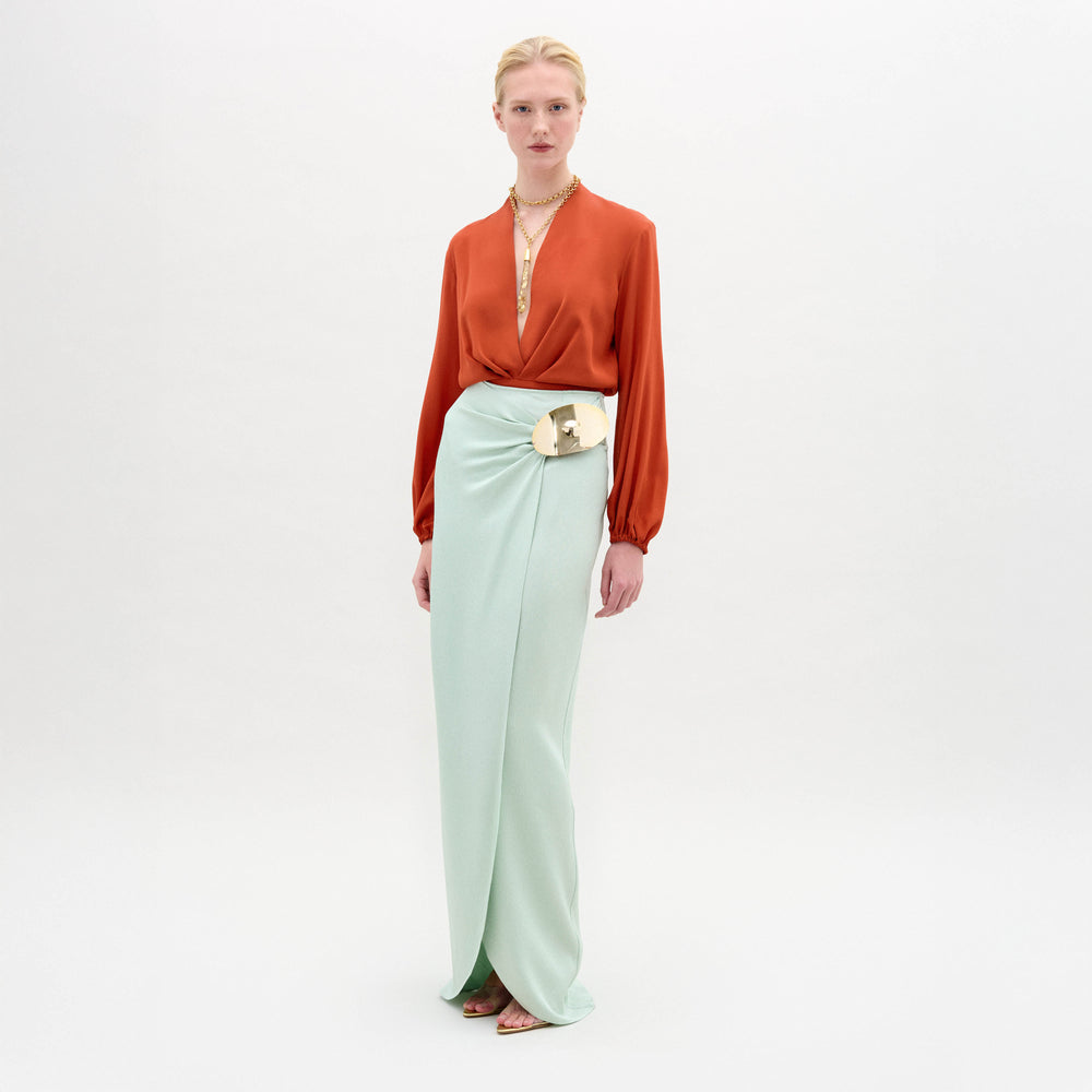 A person stands against a plain white background wearing the Gadea Skirt in light blue Italian fabric, an orange long-sleeve blouse, a gold belt, and a necklace.