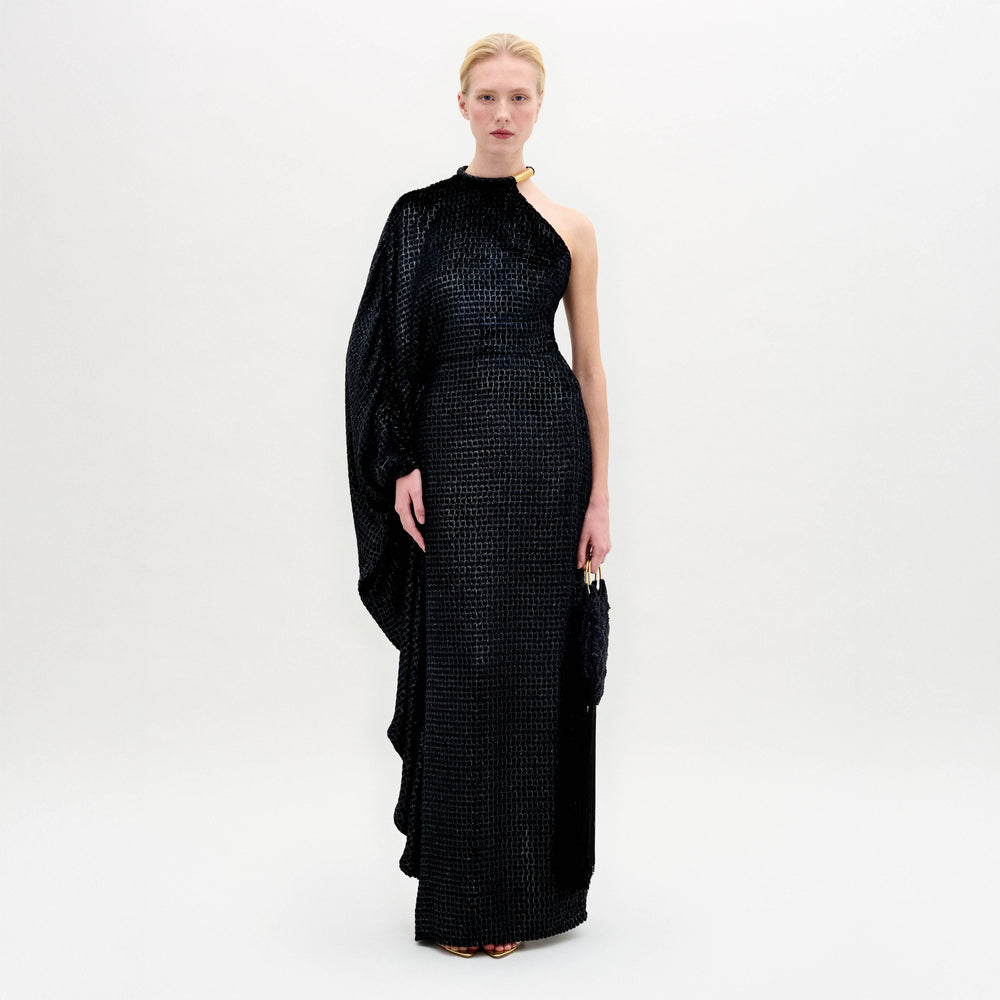 A woman stands against a plain background wearing the Gael Dress—a black, floor-length maxi with an asymmetrical neckline and textured pattern—holding a small clutch in her right hand.