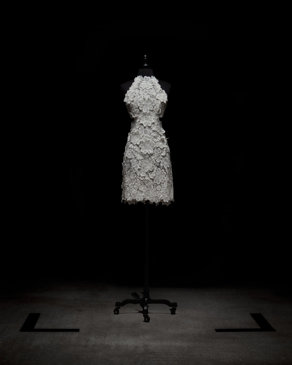 A person in the Garazi Dress, featuring sleeveless floral lace, wears sheer gloves and holds a bouquet. They pair the dress with white ankle boots while standing against a plain background.