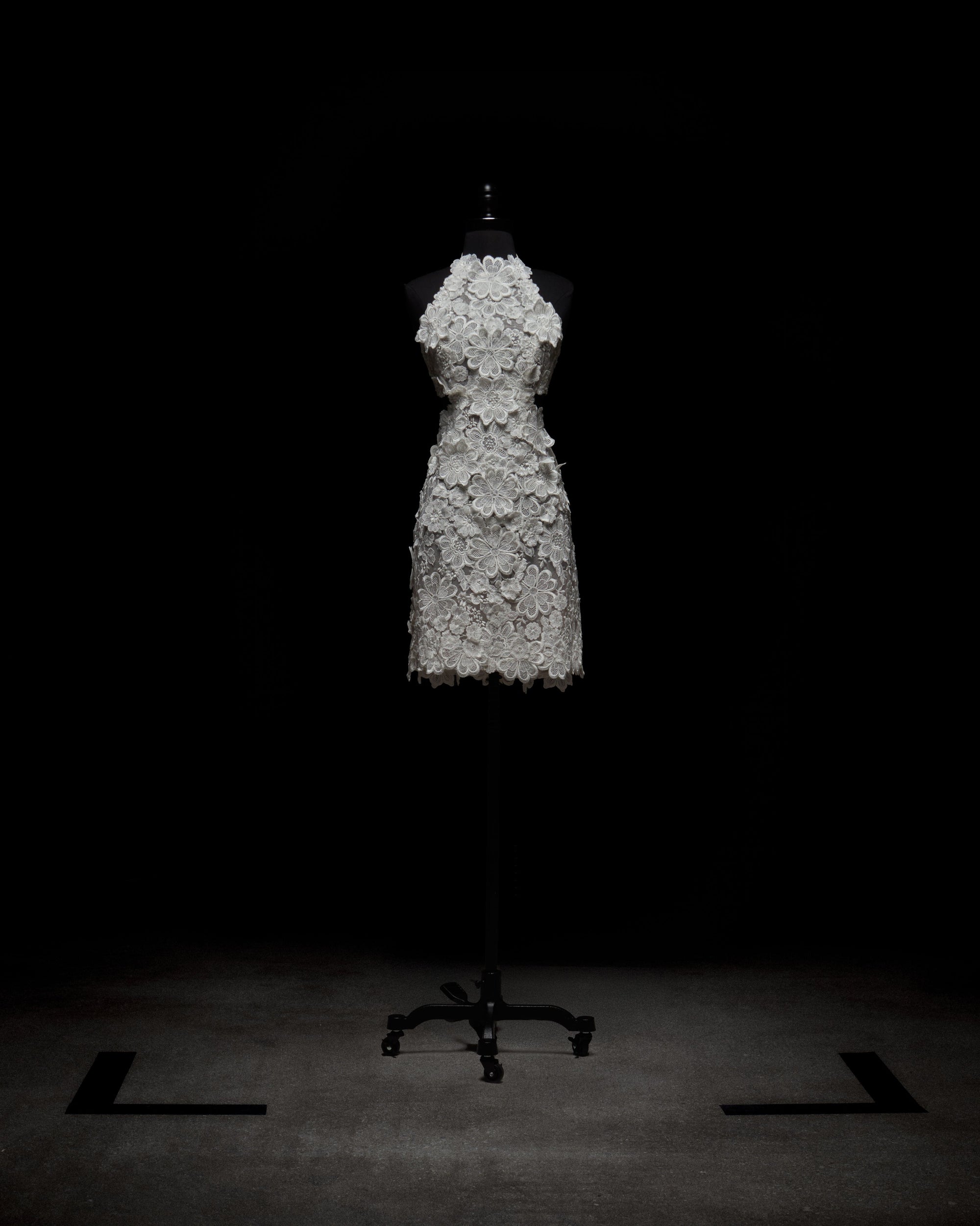 A person in the Garazi Dress, featuring sleeveless floral lace, wears sheer gloves and holds a bouquet. They pair the dress with white ankle boots while standing against a plain background.