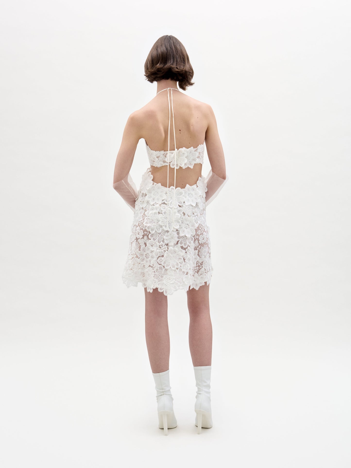 A person in the Garazi Dress, featuring sleeveless floral lace, wears sheer gloves and holds a bouquet. They pair the dress with white ankle boots while standing against a plain background.