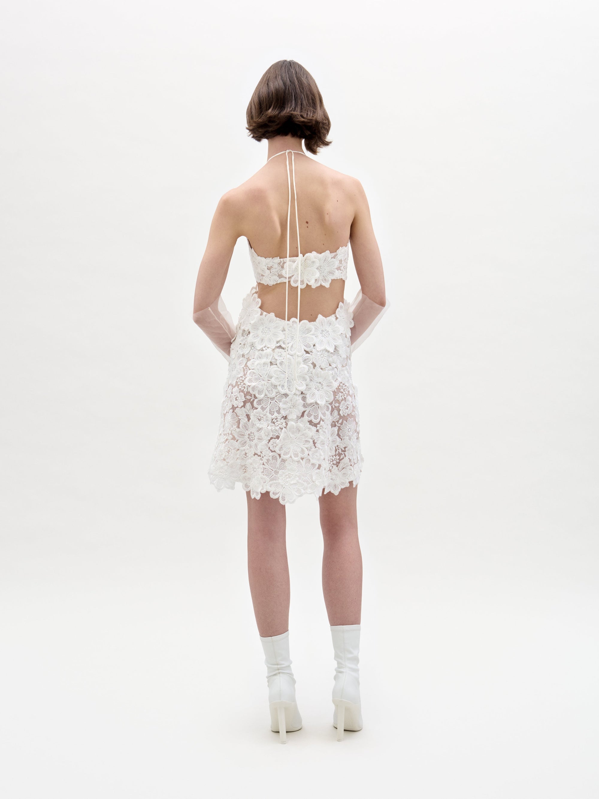 A person in the Garazi Dress, featuring sleeveless floral lace, wears sheer gloves and holds a bouquet. They pair the dress with white ankle boots while standing against a plain background.