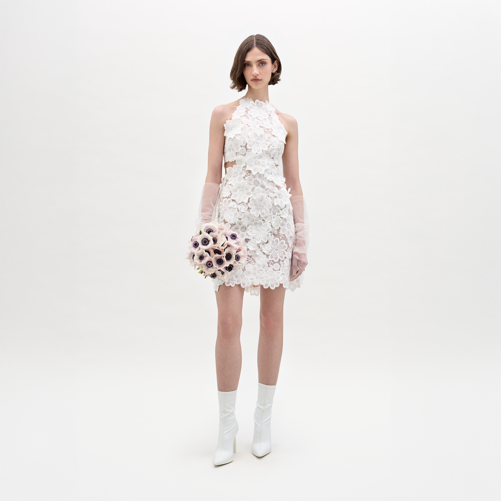 A person in the Garazi Dress, featuring sleeveless floral lace, wears sheer gloves and holds a bouquet. They pair the dress with white ankle boots while standing against a plain background.