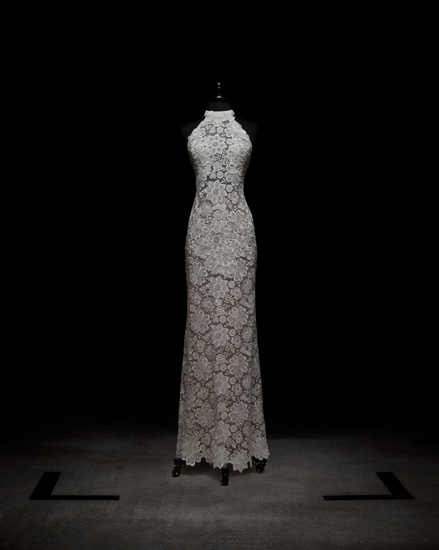 A woman in a Gardenia Dress holds pink flowers. She wears long, sheer gloves and stands against a plain background.