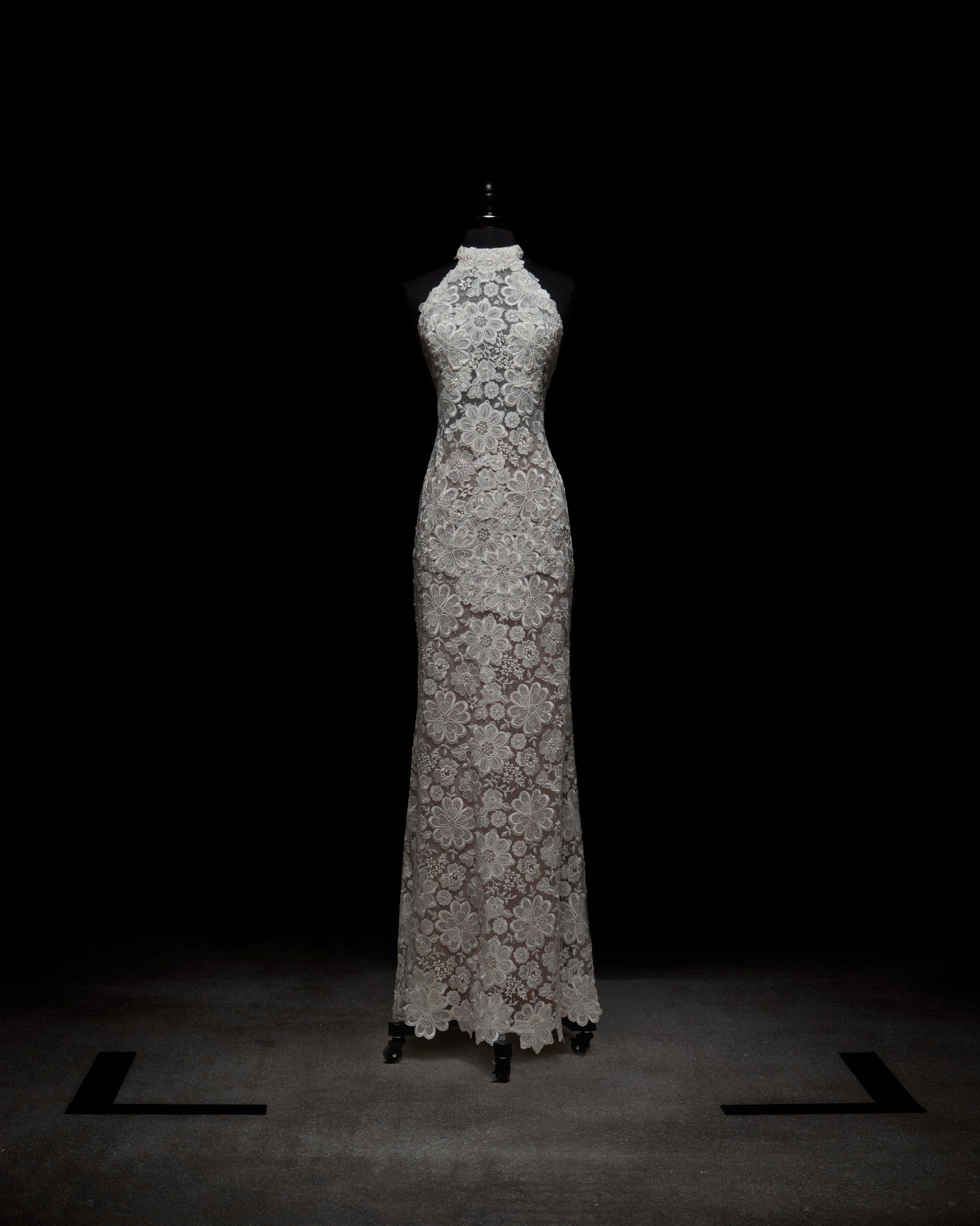 A woman in a Gardenia Dress holds pink flowers. She wears long, sheer gloves and stands against a plain background.