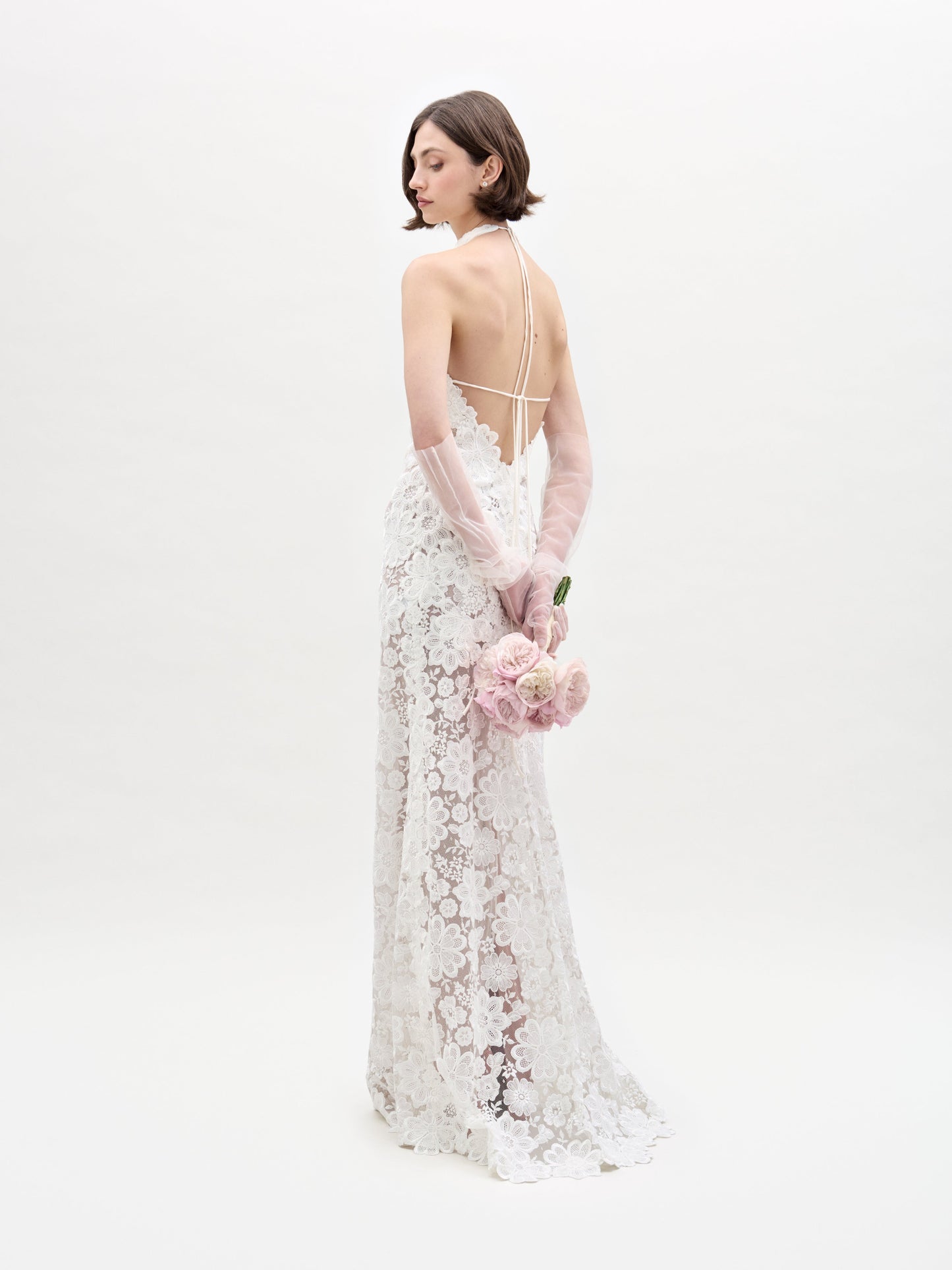 A woman in a Gardenia Dress holds pink flowers. She wears long, sheer gloves and stands against a plain background.