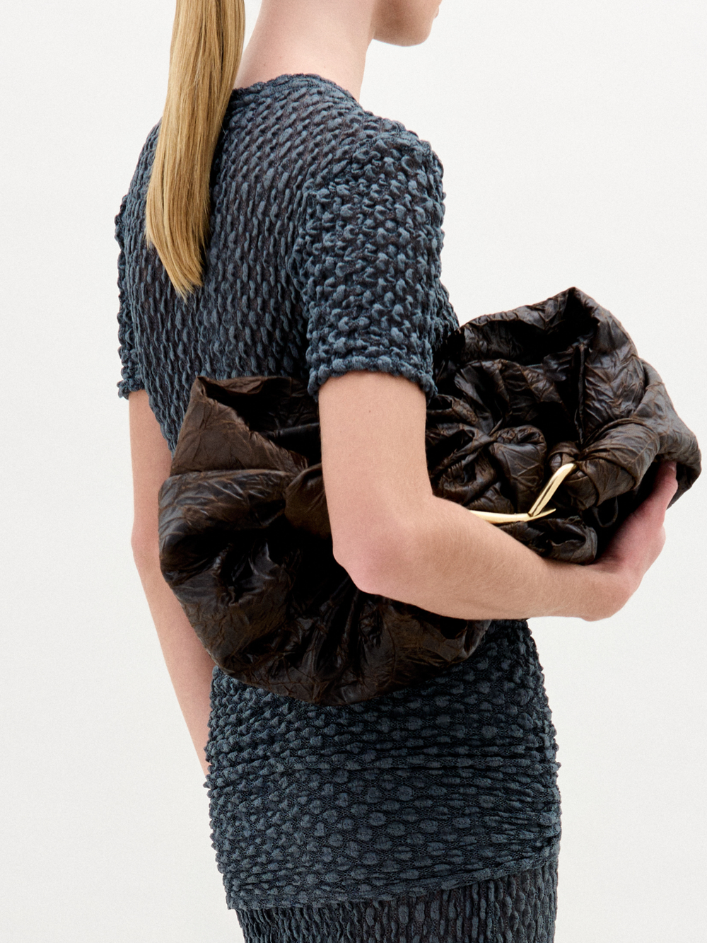 A woman with blonde hair in a low ponytail wears the Gen Tshirt, a textured, fitted, dark short-sleeve top with knot detail, and holds a large dark ruched bag against a plain white background.