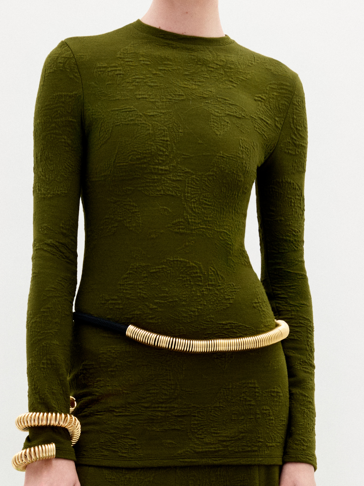 A woman stands against a plain white background wearing the Gen Tshirt, a slim fit, textured olive green long-sleeve dress with gold accessories and holding a brown bag.