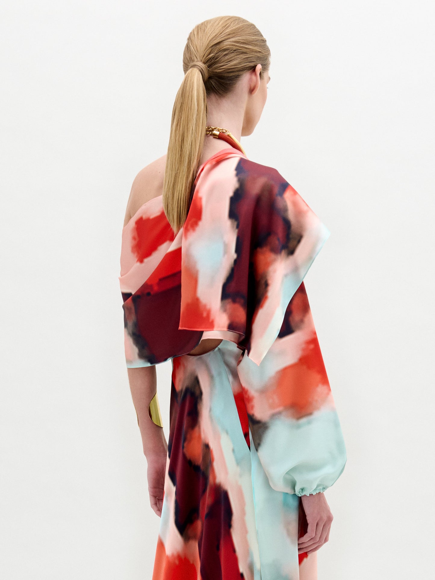 A woman stands against a plain white background wearing the Gery Shrug, a long-sleeved dress featuring a red, blue, and white abstract pattern on Italian fabric, paired with a large gold collar-style necklace.