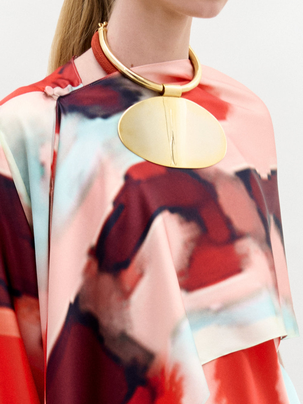 A woman in a colorful abstract-patterned dress accessorized with a circular gold necklace and the Gery Shrug stands against a plain background.