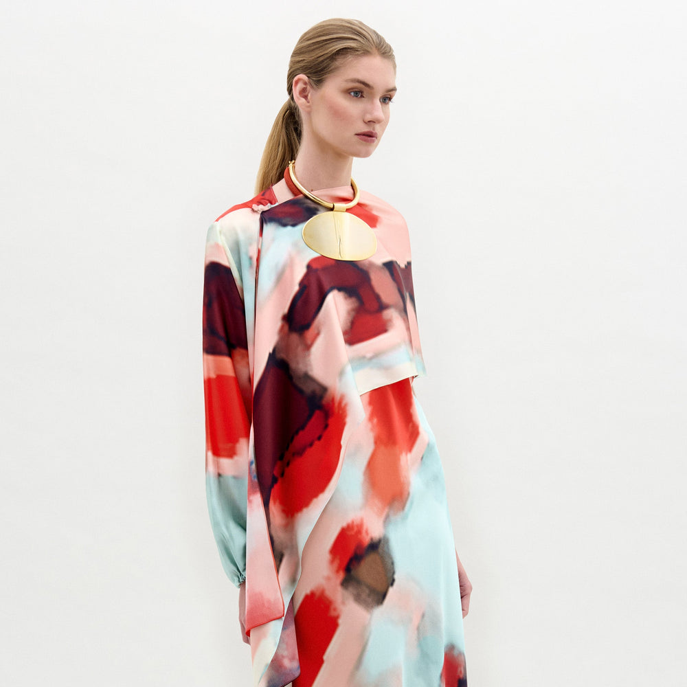 A woman in a colorful abstract-patterned dress accessorized with a circular gold necklace and the Gery Shrug stands against a plain background.