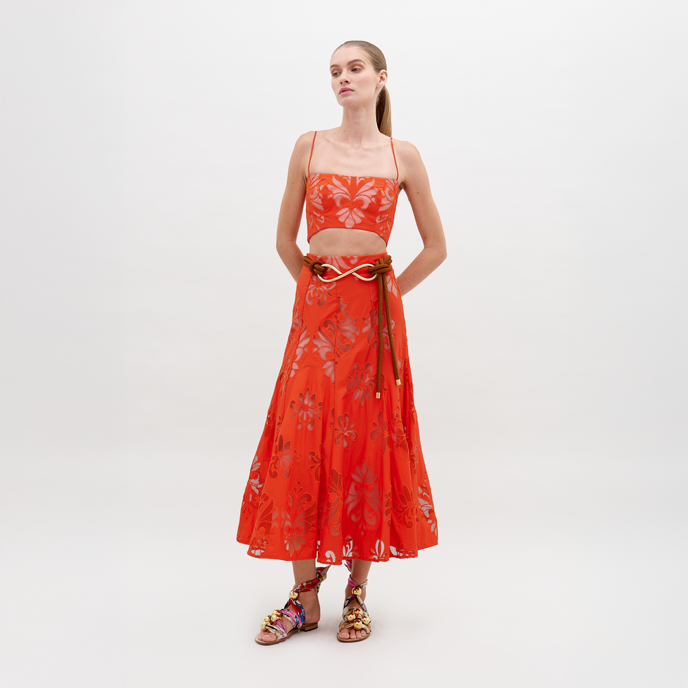 A woman stands wearing a Grettel Skirt Rouge Embroidery with a bright orange floral-patterned two-piece outfit and colorful sandals. She poses against a plain white background, available for pre-order starting November 15th for the 2024 collection.