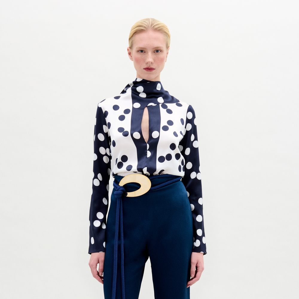 A woman with pale skin and blonde hair wears the Gaby Blouse—a navy and white polka dot silk top with a keyhole neckline—paired with blue pants and a large circular belt buckle, standing against a plain background.
