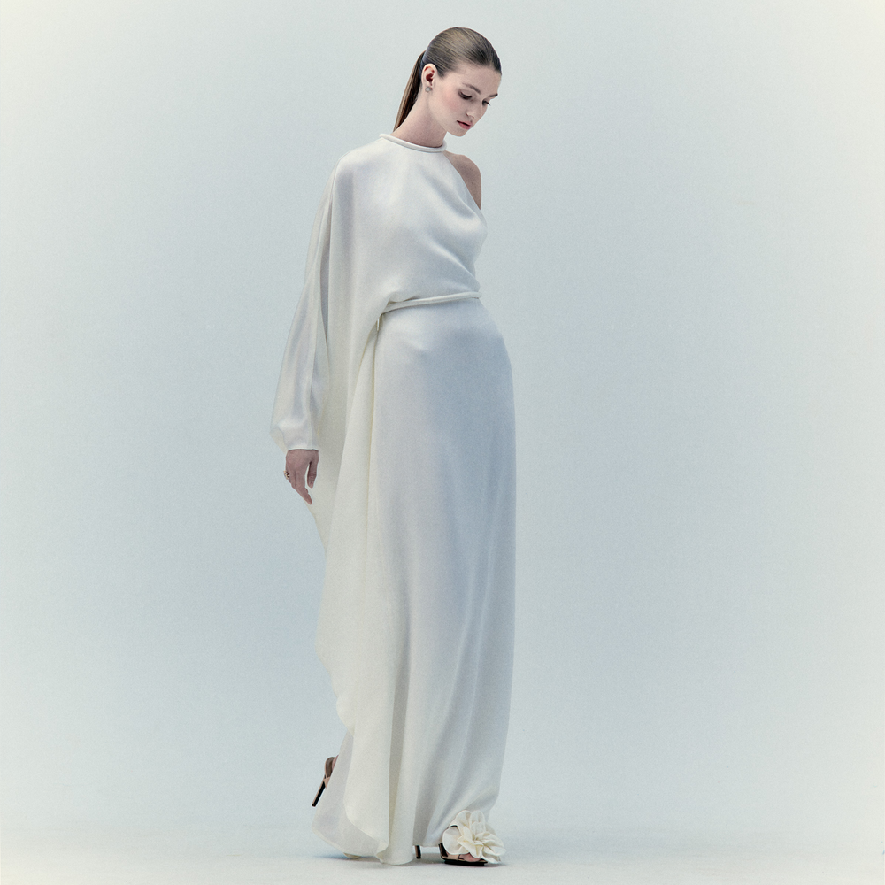 A woman wears the Gael Dress White— a long maxi dress with an asymmetric neckline—standing against a light background, looking down with her hair tied back.