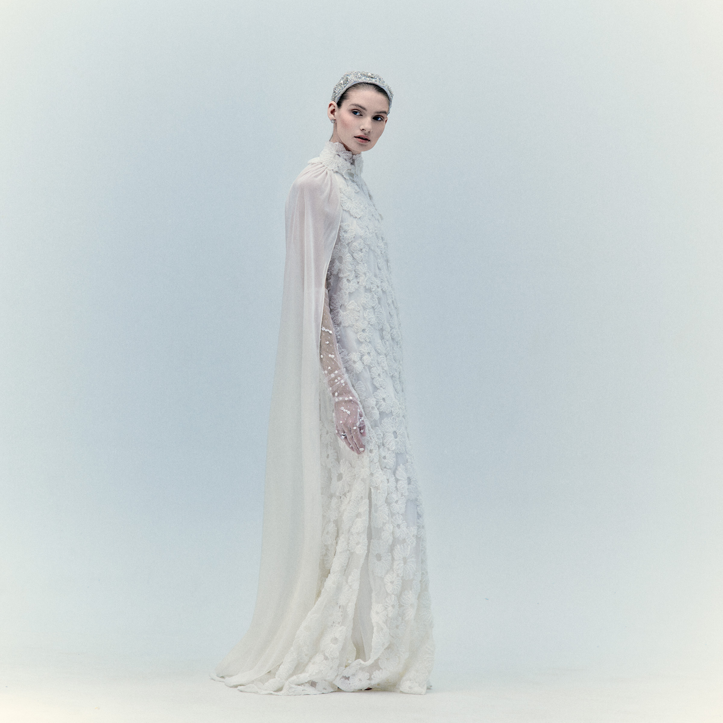 A woman in the Gertrude Dress Pearl, featuring intricate floral appliqué, sheer sleeves, and a matching headpiece, stands against a plain, light background.