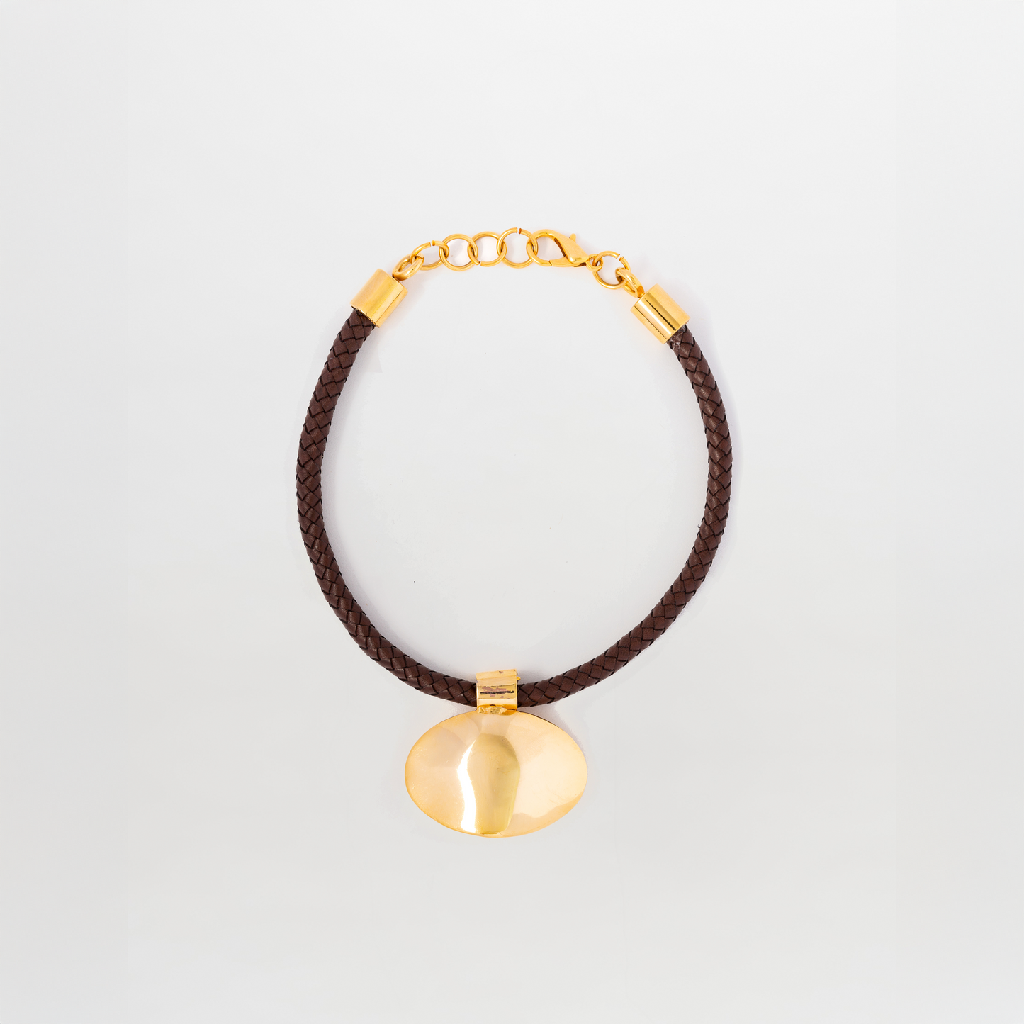The Gimena Necklace features a gold disc pendant on a dark brown braided cord with gold accents and an adjustable chain clasp. Made in Colombia.
