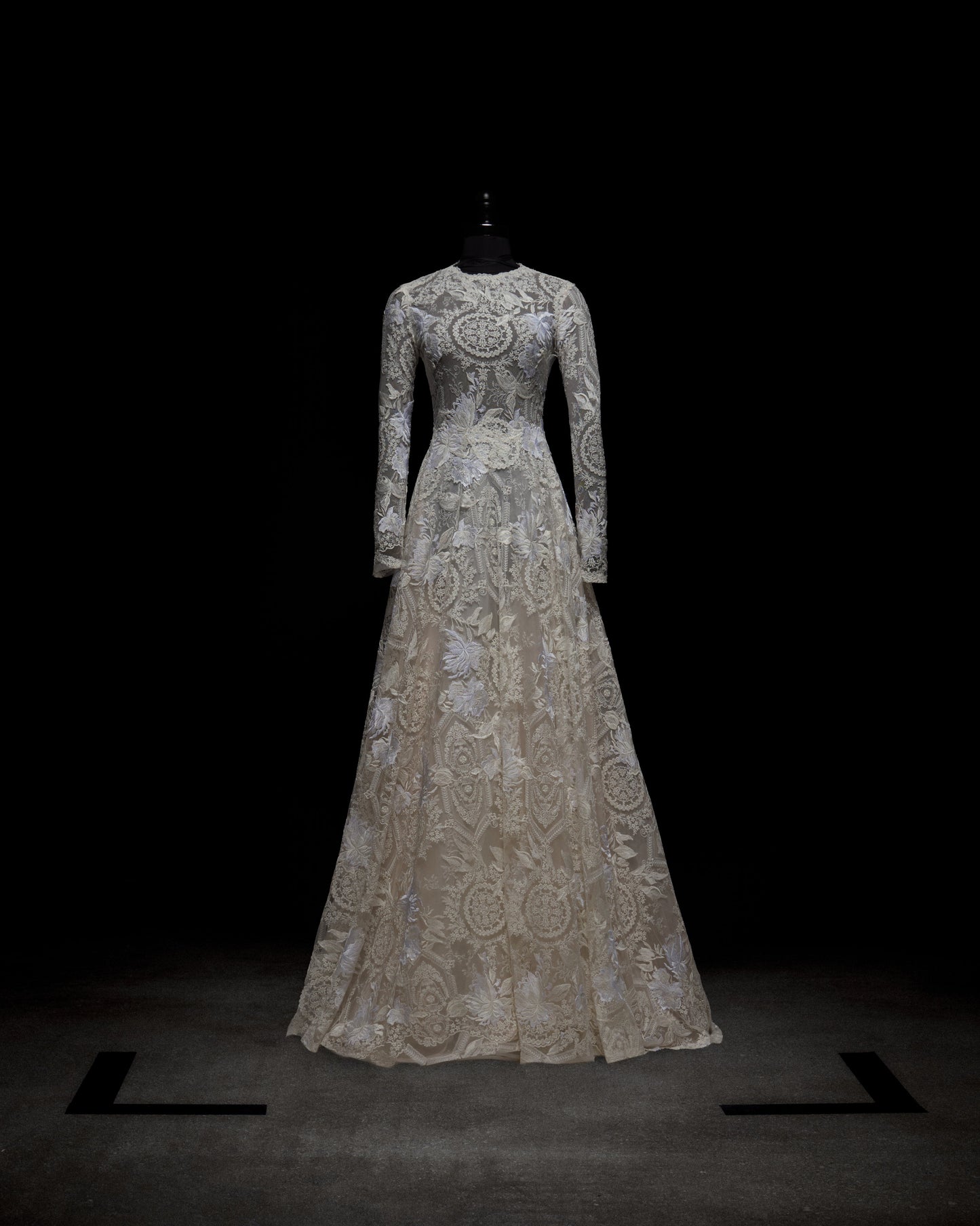 A woman poses against a plain background in the Hermoine Dress, an ornate lace bridal gown featuring long sleeves and a flowing train.