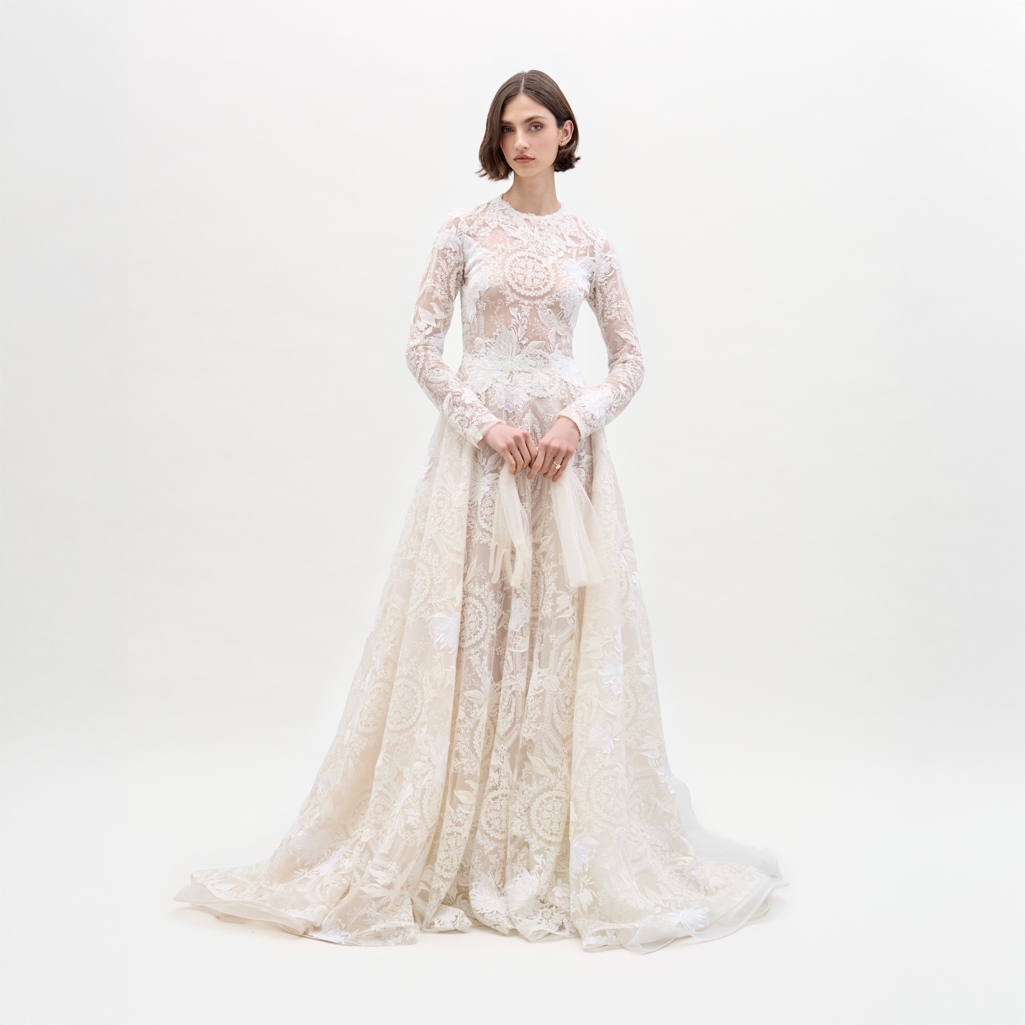 A woman poses against a plain background in the Hermoine Dress, an ornate lace bridal gown featuring long sleeves and a flowing train.