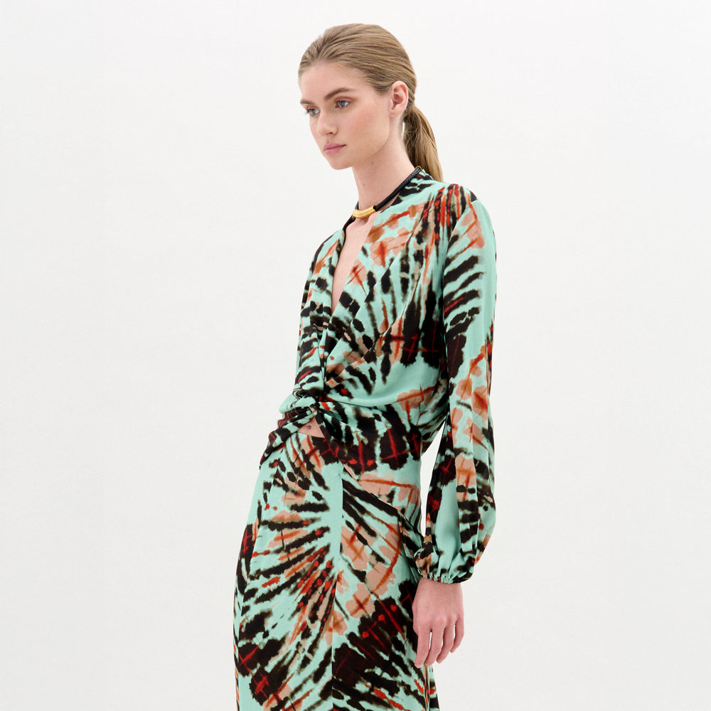 A woman stands against a plain white background wearing the Honey Blouse, a long-sleeved top featuring green, black, and orange abstract patterns with a chic front twist detail.