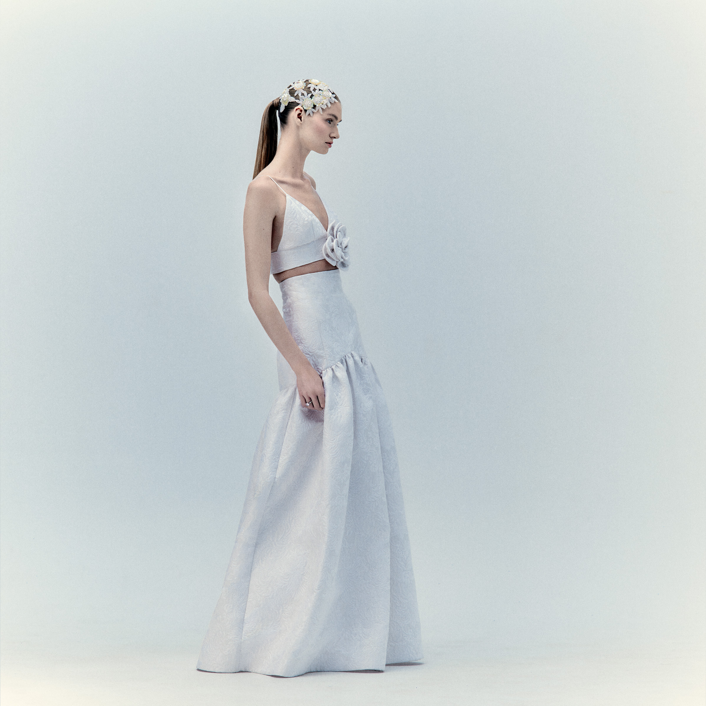 A woman stands in profile wearing the Helen Set Dress Pearl, a white two-piece A-line outfit with 3D flower appliqué and a decorative headpiece, set against a plain, light background.