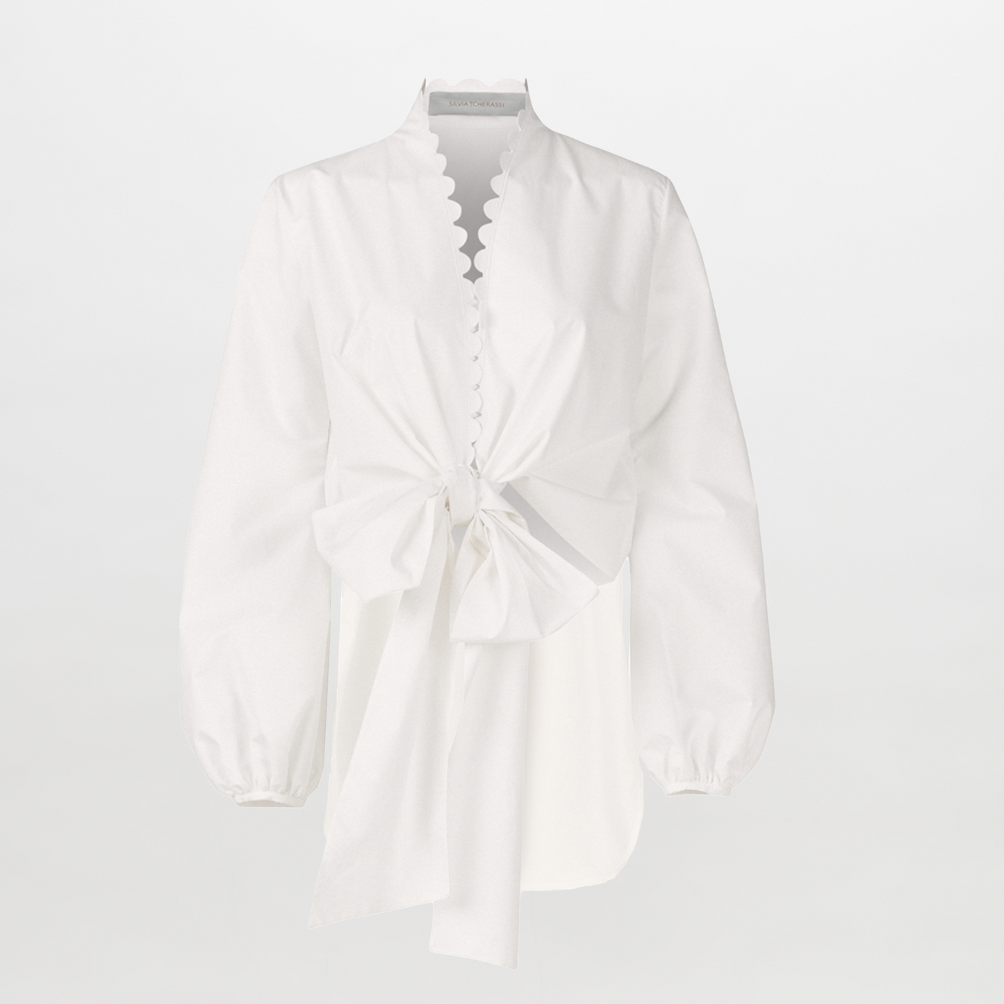The Honey Blouse White Eyelet features white cotton with organza sleeves, scalloped edges, a distinctive neckline, front bow detail, and an asymmetrical hem, shown against a plain white background.