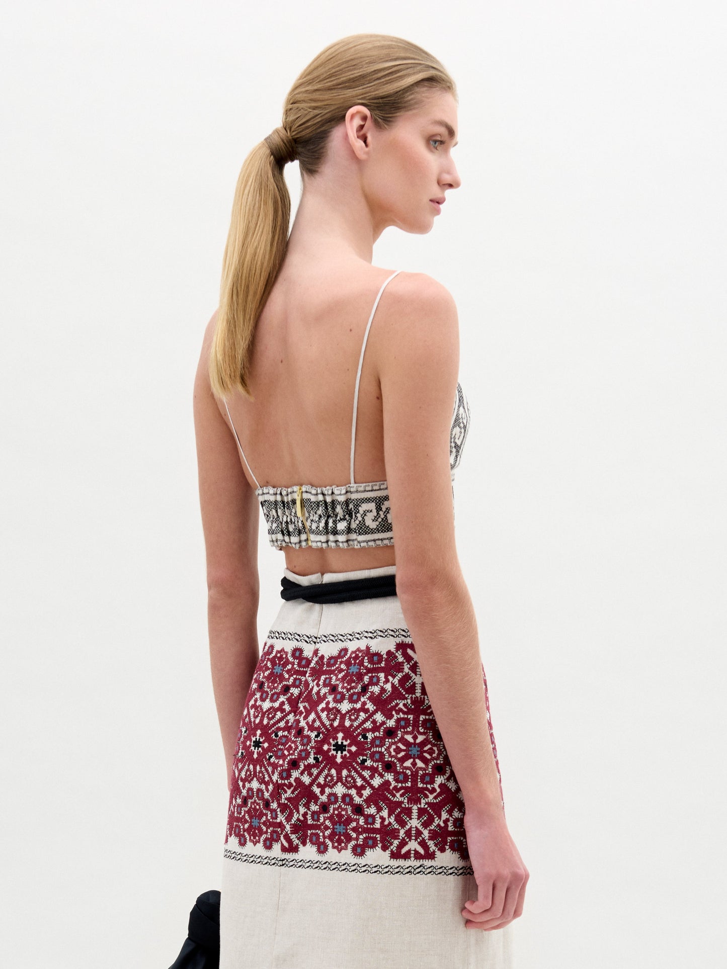 A woman in the Ibbie Top, a sleeveless V-neck with geometric patterns, paired with a red and white Italian-fabric skirt and a large decorative belt, stands against a plain background.
