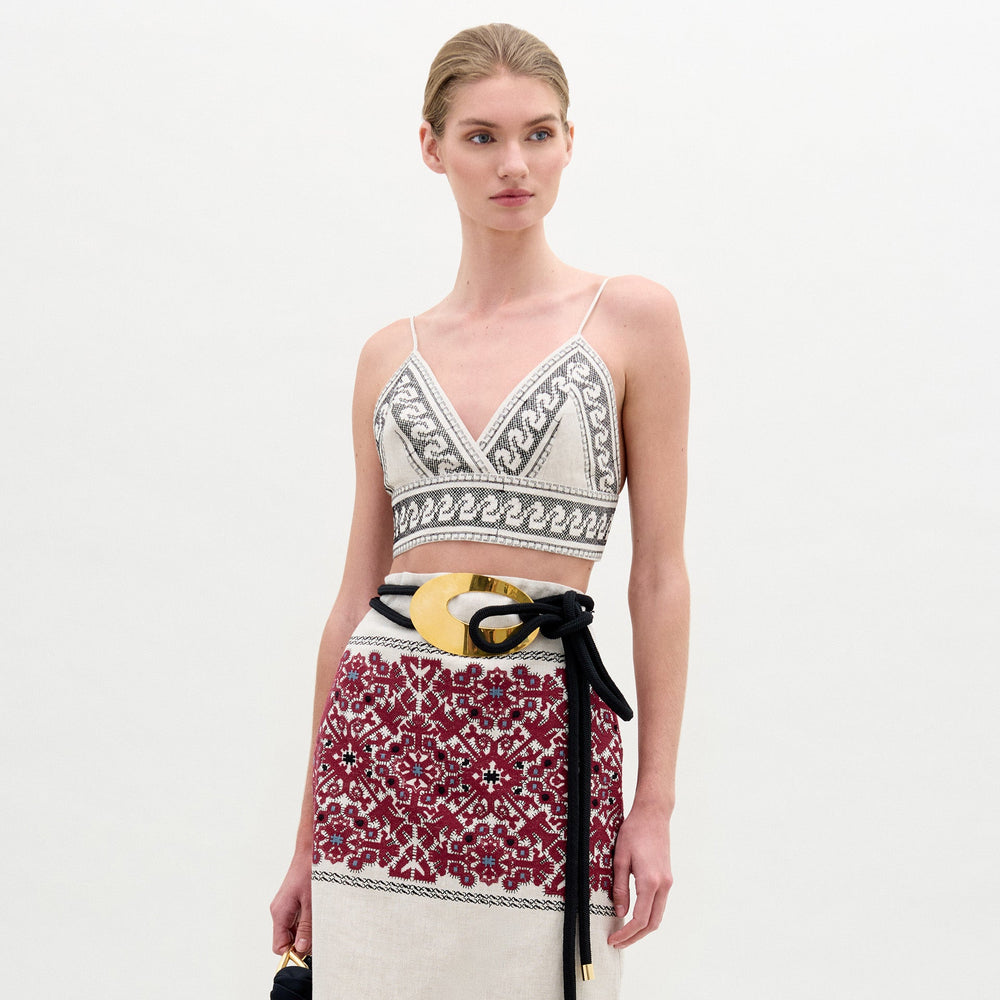 A woman in the Ibbie Top, a sleeveless V-neck with geometric patterns, paired with a red and white Italian-fabric skirt and a large decorative belt, stands against a plain background.
