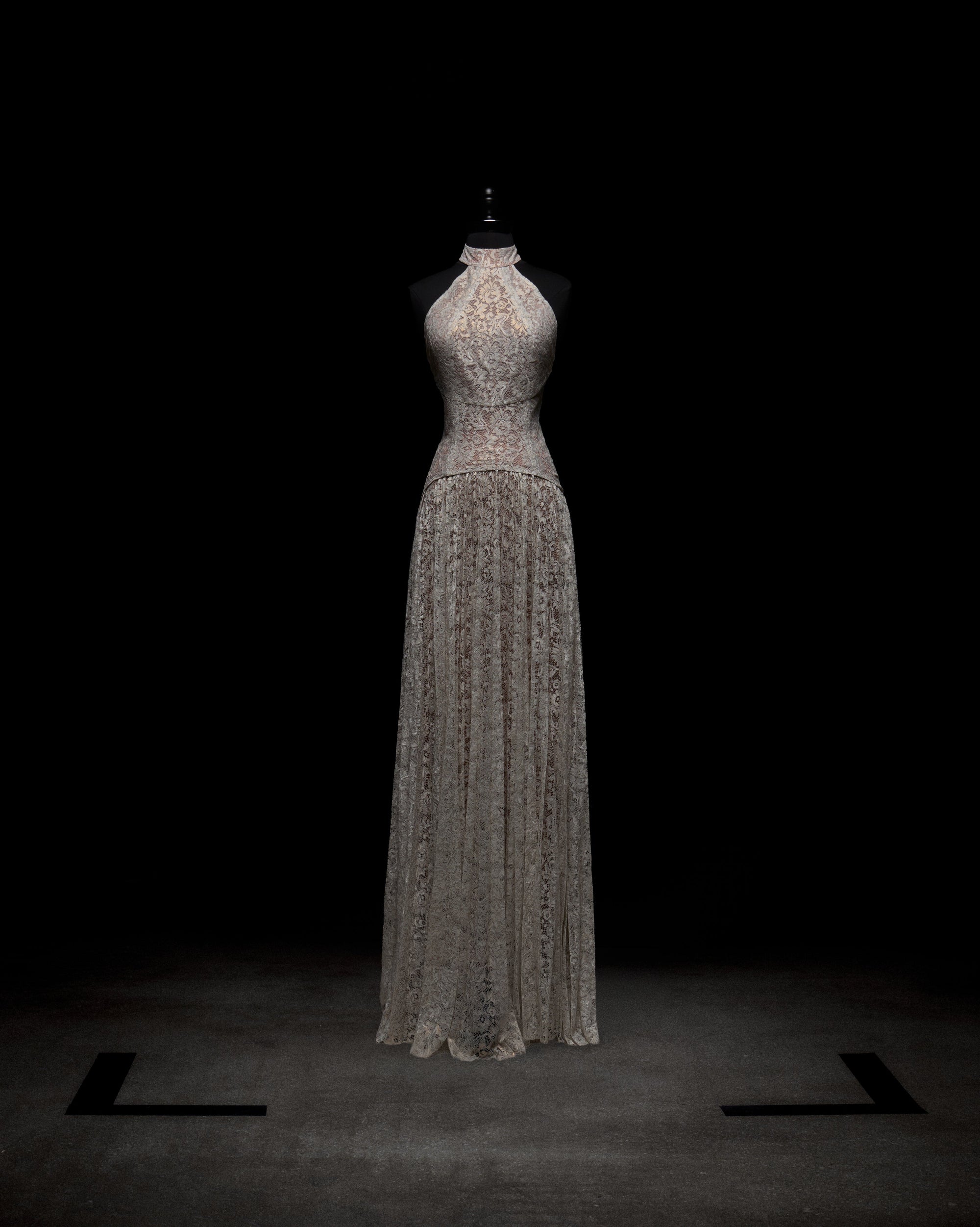 Wearing the Ilana Dress, a woman in a long lace bridal gown holds a bouquet of white and purple flowers against a plain background.