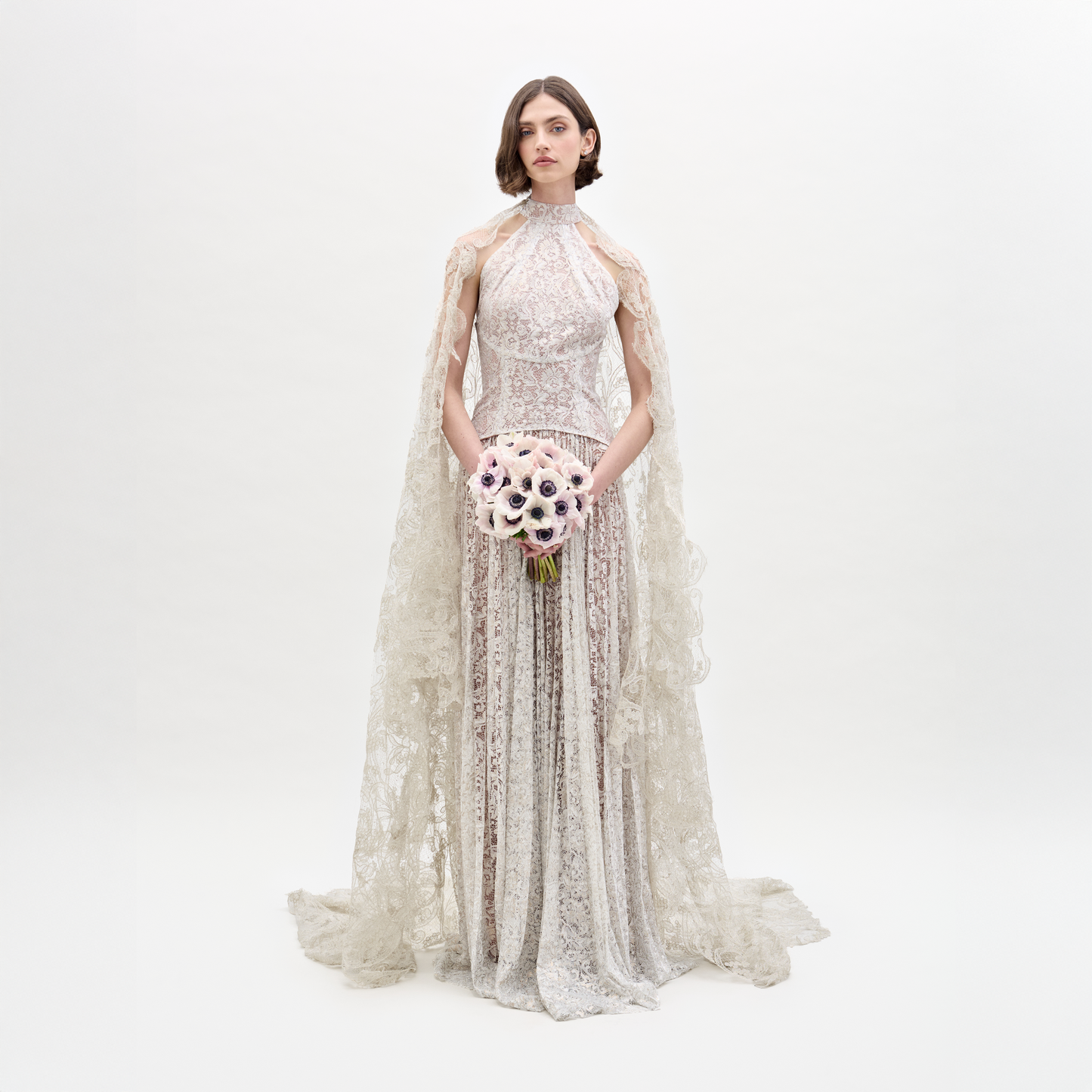 Wearing the Ilana Dress, a woman in a long lace bridal gown holds a bouquet of white and purple flowers against a plain background.