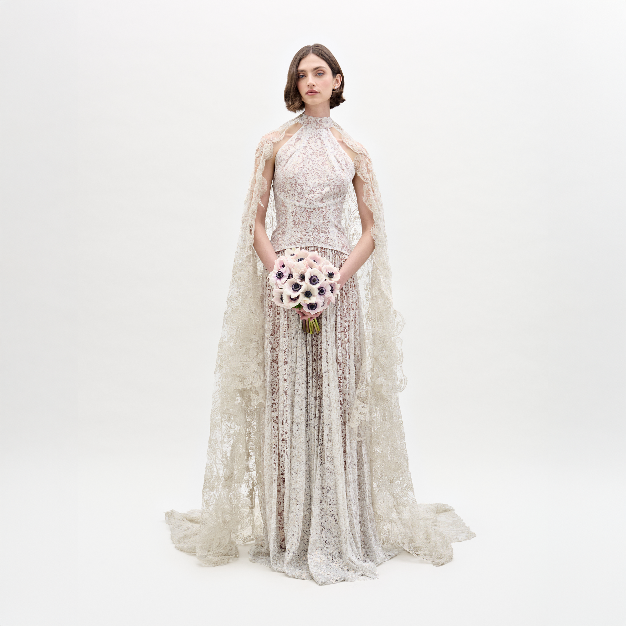 Wearing the Ilana Dress, a woman in a long lace bridal gown holds a bouquet of white and purple flowers against a plain background.